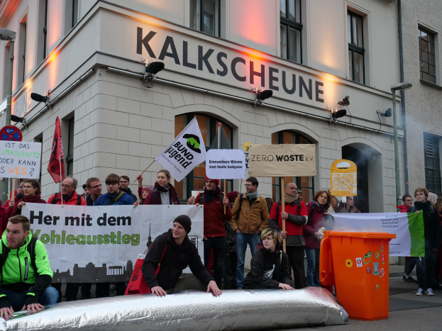 Eine Gruppe von Menschen mit Protestschildern steht vor einem Gebäude mit Fenstern und Schildern, während zwei Personen im Vordergrund auf einem Objekt sitzen und ein Müllcontainer rechts zu sehen ist.