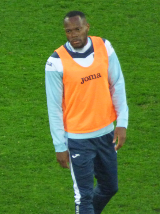 Ein Füßballer in einem orangen T-Shirt und blauen Hosen überquert entschlossen ein grünes Feld.