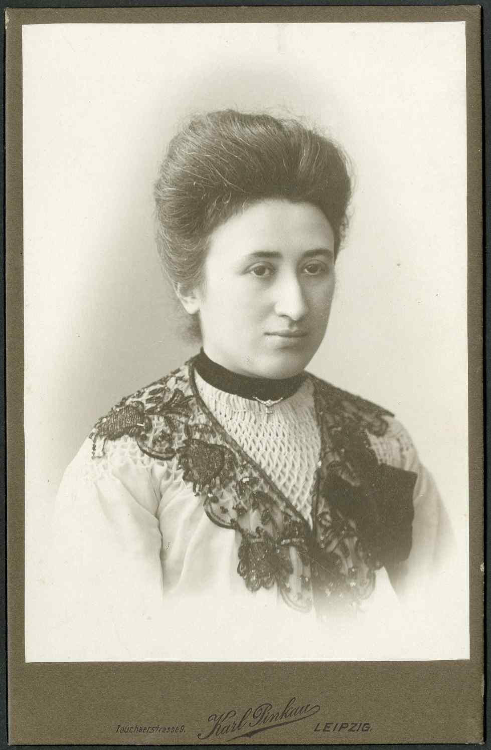 Schwarzes und weißes Foto einer Frau in einem weißen Kleid mit einem gemusterten Schultertuch, identifiziert als Karl Pinkau Leipzig, die direkt in die Kamera schaut.