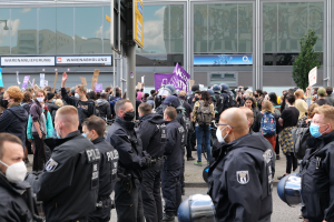Eine große Gruppe von Menschen steht vor einem Gebäude, einige halten Schilder und tragen Helme, im Vordergrund ein Mast mit einer Tafel und im Hintergrund ein Baum, sie scheinen zu protestieren.