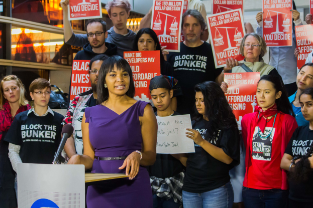 Eine Black-Lives-Matter-Aktivistin steht an einem Rednerpult mit einem Mikrofon, spricht zu einer Menge mit Schildern, mit Lichtern und Gegenständen im Hintergrund.