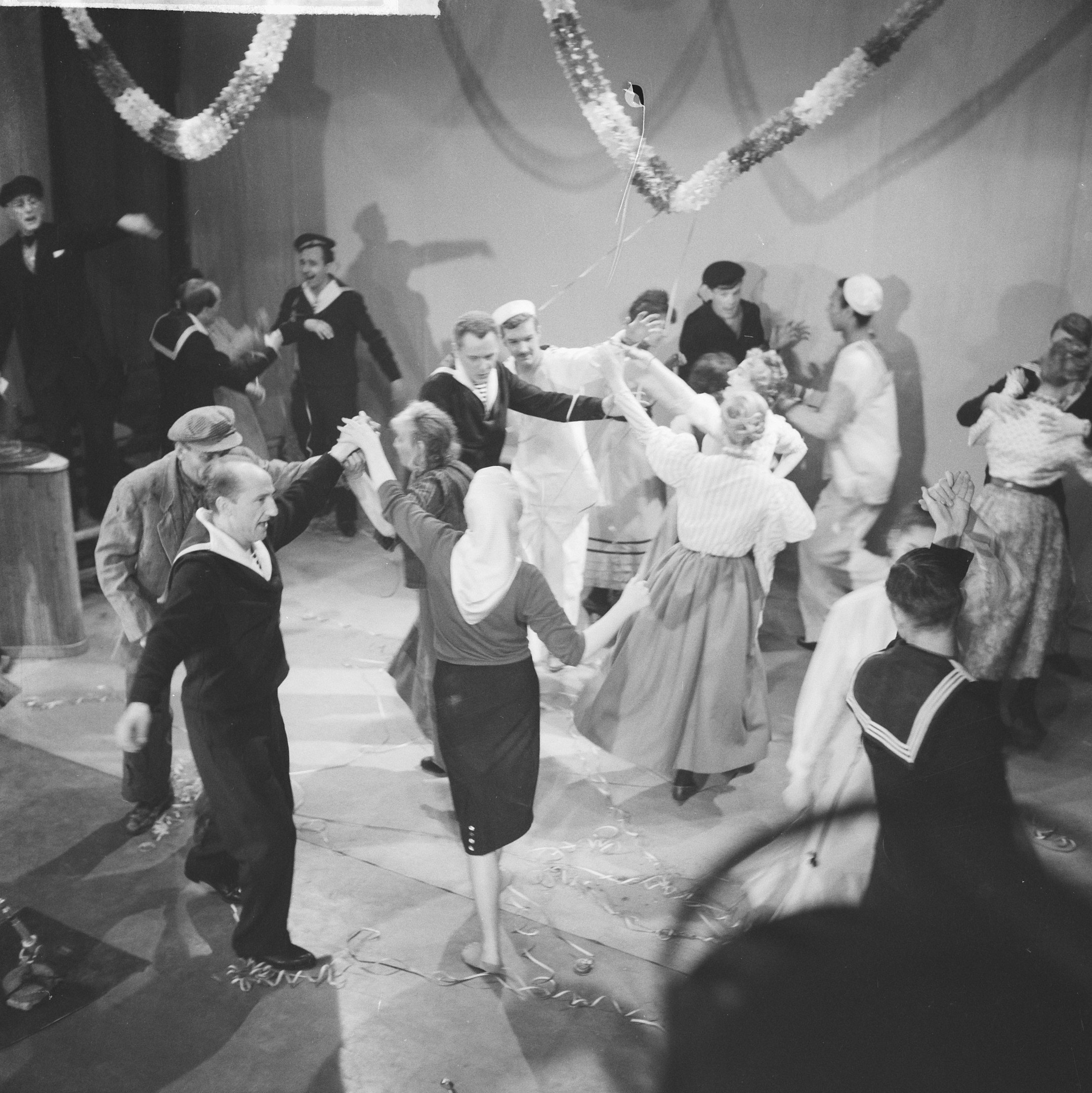 Schwarz-weiß-Bild von Menschen, die auf einer Tanzfläche tanzen, mit einer dekorierten Wand dahinter und einem Tisch mit Gegenständen links.
