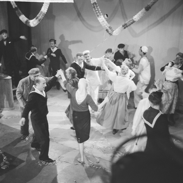 Schwarz-weiß-Bild von Menschen, die auf einer Tanzfläche tanzen, mit einer dekorierten Wand dahinter und einem Tisch mit Gegenständen links.