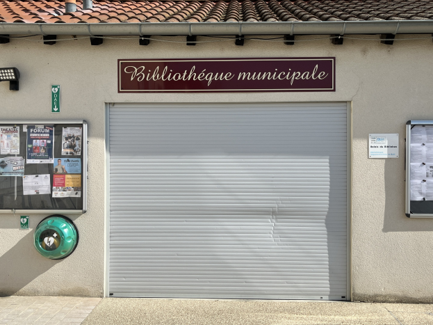Ein Rathausgebäude mit der Aufschrift 'Bibliothèque municipale', mit einem zentralen Rollladen, einer Texttafel, Plakaten an der linken Wand und einem beleuchteten Weg zur Eingangstür.