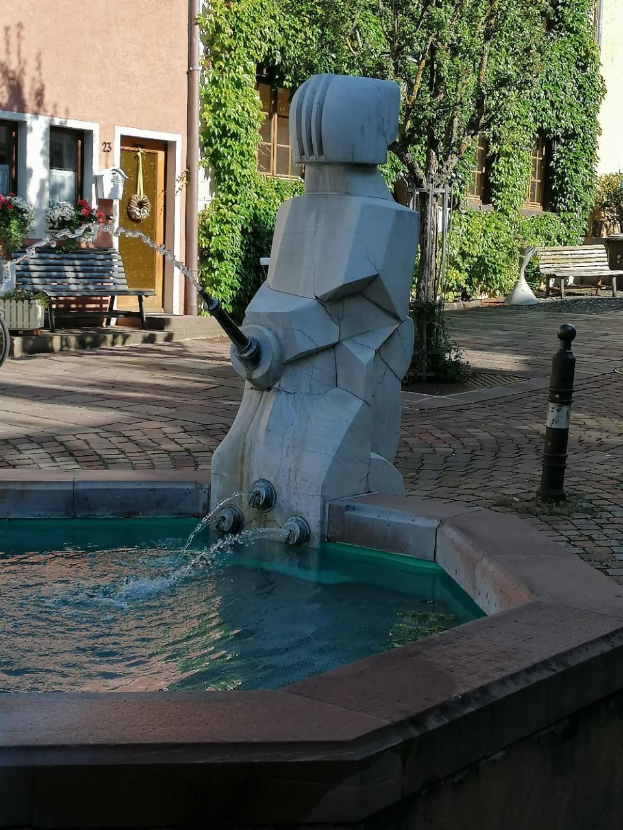 Ein Stadtplatzbrunnen umgeben von Gebäuden, Grünflächen, Bänken und Blumenkübeln, mit Wasser, das eine friedliche Szenerie schafft.