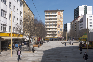 Ein belebter Stadtplatz mit Fußgängern, Bänken, Bäumen, Gebäuden, einem Karussell und Oberleitungen.