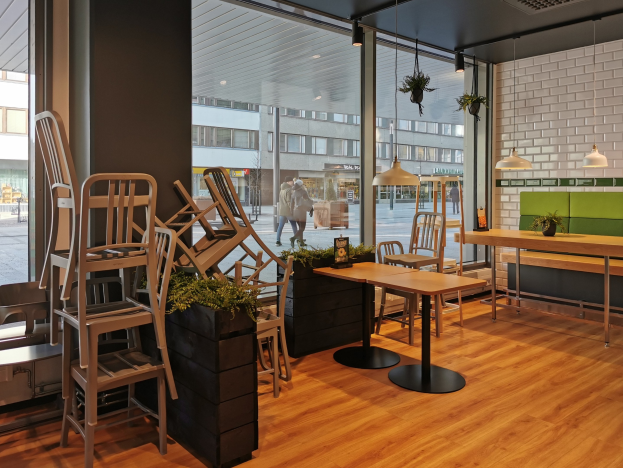 Ein Restaurant-Innenraum mit Holztischen und -stühlen in der Nähe eines großen Fensters, das auf eine Straße in Berlin mit Passanten, Gebäuden und urbanen Elementen blickt.