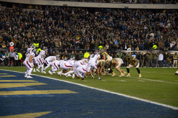 Ein Fussballspiel im Gange mit Spielern in Sportkleidung und Helmen auf dem Feld, Zuschauern, Gelöndern und Tafeln mit Text im Hintergrund.