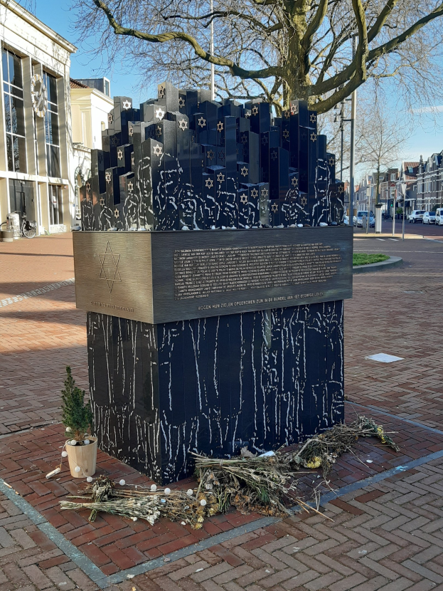 Ein Holocaust-Mahnmal in einem Stadtplatz, mit einem Baum davor, einem Topfgewächs auf dem Boden und verstreuten trockenen Blättern, mit Gebäuden, Fahrzeugen, Pfosten und Bäumen im Hintergrund.