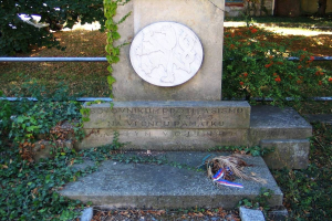 Ein Gedenkmonument auf einem Friedhof, umgeben von einem Geländer, Pflanzen, Blumen und Bäumen, mit einem Kranz darauf, gewidmet den Opfern des Zweiten Weltkriegs, vor einem Gebäude mit Fenstern und einem Boden, der mit trockenen Blättern und Gras bedeckt ist.