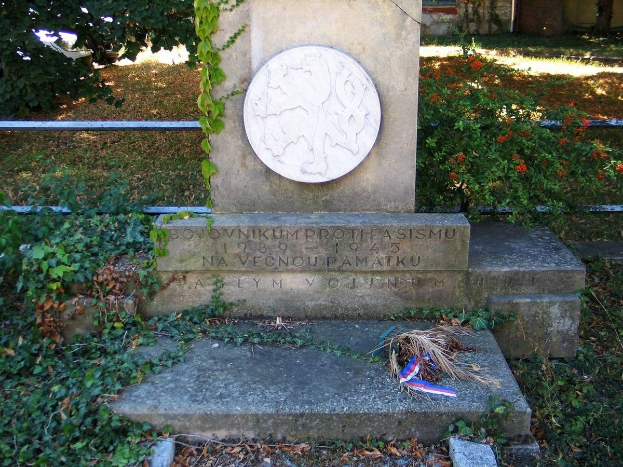 Ein Gedenkmonument auf einem Friedhof, umgeben von einem Geländer, Pflanzen, Blumen und Bäumen, mit einem Kranz darauf, gewidmet den Opfern des Zweiten Weltkriegs, vor einem Gebäude mit Fenstern und einem Boden, der mit trockenen Blättern und Gras bedeckt ist.