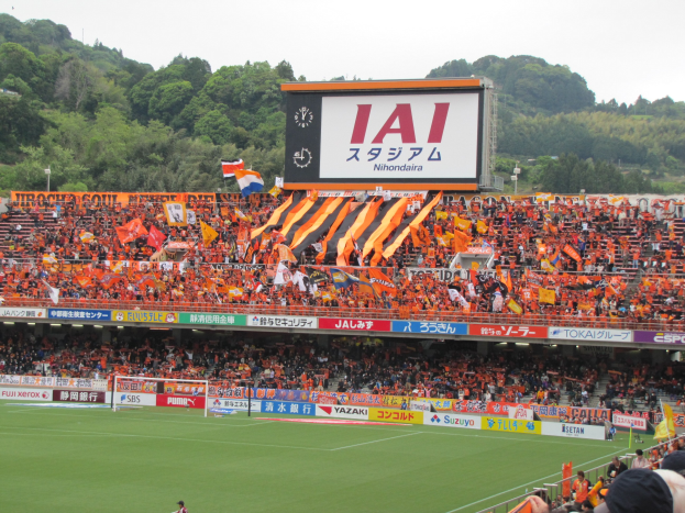 Fußballspiel in einem Stadion mit großer Zuschauermenge, saftig grünem Rasen, einem Tor, Schildern, Fahnen, einem großen Bildschirm, Bäumen und einem klaren blauen Himmel.