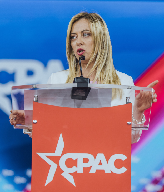 Frau in einem weißen Kleid, die an einem Rednerpult mit einem Mikrofon spricht und wie eine politische Wissenschaftlerin aussieht.