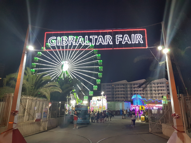 Der Eingang zur Gibraltar-Messe bei Nacht, mit einem hell erleuchteten Riesenrad, einer leuchtenden Namensanzeige, Menschenmassen, Bäumen und Gebäuden im Hintergrund unter einem dunklen Himmel.