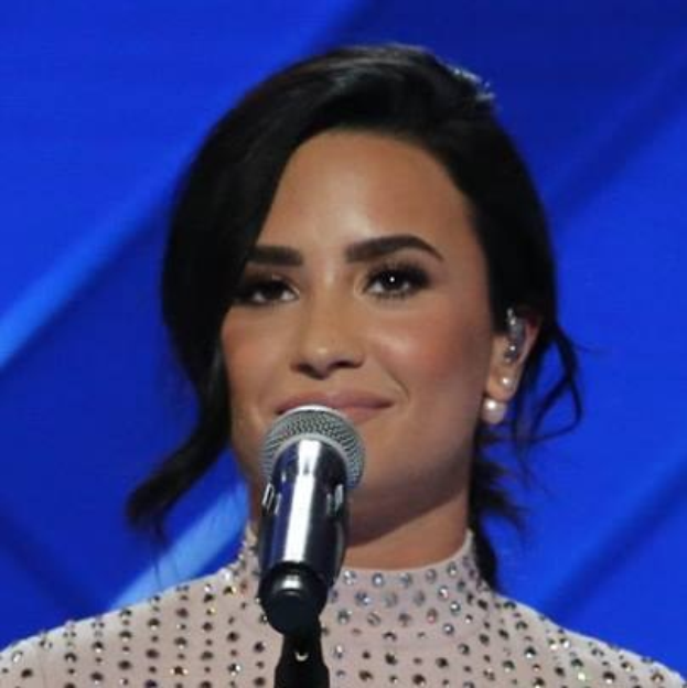 Demi Lovato bei den Grammys in einem schwarzen Kleid und einem eleganten Bob, selbstbewusst vor einem Mikrofon mit blauem Hintergrund stehend.