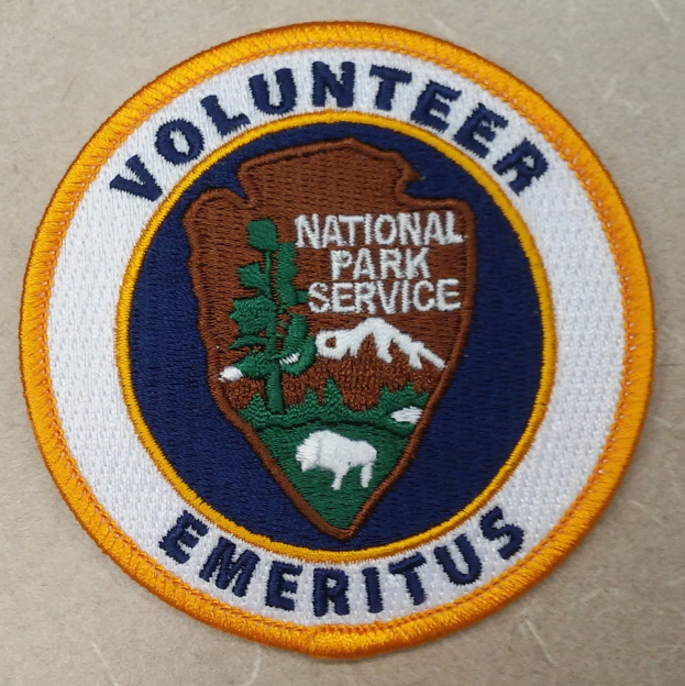 Ein kreisföriges Abzeichen mit einem blauen Rand, weißem Hintergrund und fetter schwarzer Schrift, die 'Volunteer National Park Service Emeritus' in der Mitte eines gelben Kreises lesen.