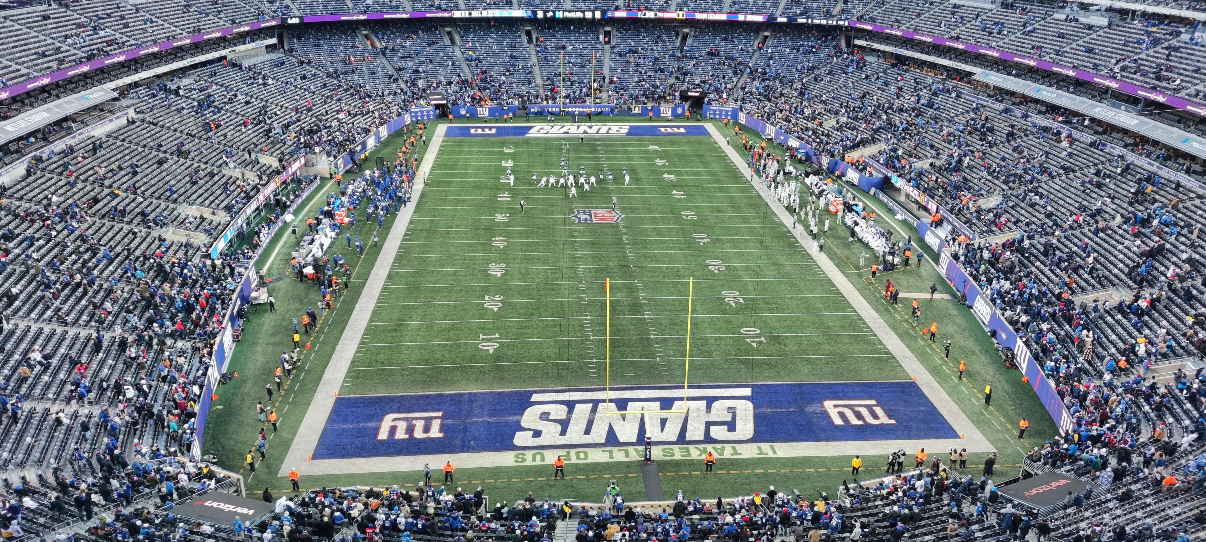 Ein voller Fußballstadion mit Spielern auf dem Feld, Torpfosten und Texttafeln, aus einer erhöhten Perspektive betrachtet.