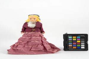 Eine Barbie-Puppe in einem rosafarbenen Kleid und einer maronenfarbenen Jacke steht neben einem schwarzen Farbpaletten auf einer weißen Oberfläche.