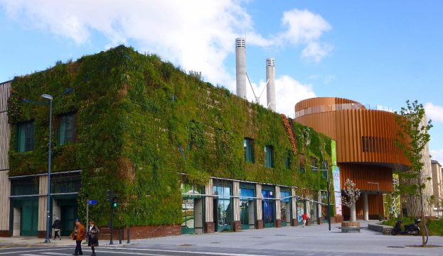 Großes nachhaltiges Gebäude mit grüner Wand, umgeben von Straßeninfrastruktur, Fahrzeugen, Fußgängern, Bäumen und bewölktem Himmel.