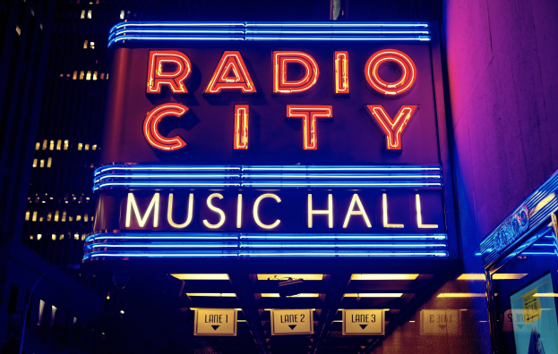 Radio City Music Hall in New York City, hell erleuchtet in der Nacht, umgeben von hohen Gebäuden und einem 'Radio City'-Schild auf der rechten Seite mit mehreren kleineren Schildern darunter.
