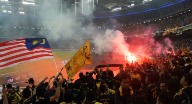 Große Menschenmenge in einem Stadion mit Feuerwerk, Fahnen, Rauch und Schildern mit der Aufschrift "Malaysia vs Malaysia Live Streaming" bei einem Ereignis.