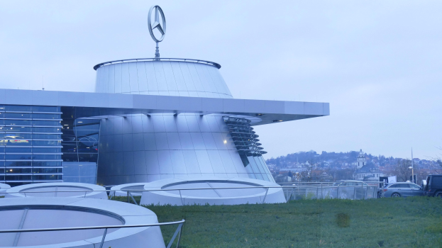 Großes modernes Mercedes-Benz-Museum-Gebäude mit Logo oben drauf, in einer parkähnlichen Anlage mit Gras, Geländern, Fahrzeugen auf der Straße, Bäumen im Hintergrund und einem klaren blauen Himmel.