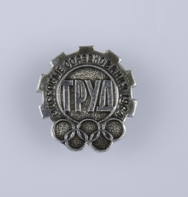 Silbernes Olympisches Abzeichen mit 'Olympische Spiele' Text auf weißem Hintergrund.