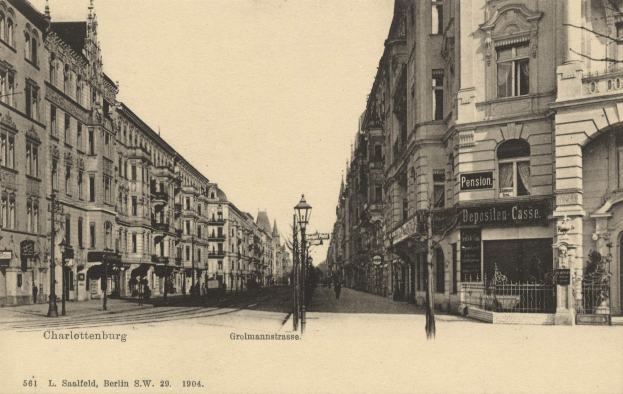 Schwarz-weiß-Fotografie einer Straßenansicht in Charlottenburg, Deutschland, mit Gebäuden, Straßenlaternen, Schildern, Motorfahrzeugen, Fußgängern, Gittern und einem klaren Himmel.