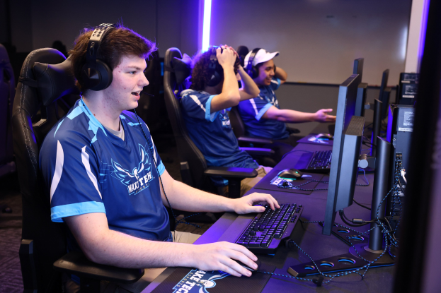 Eine Gruppe junger Männer mit Headsets spielt ein Videospiel an Computern auf einem Schreibtisch mit Monitoren, Tastaturen und anderen Gegenständen sowie beleuchteten Wänden im Hintergrund.