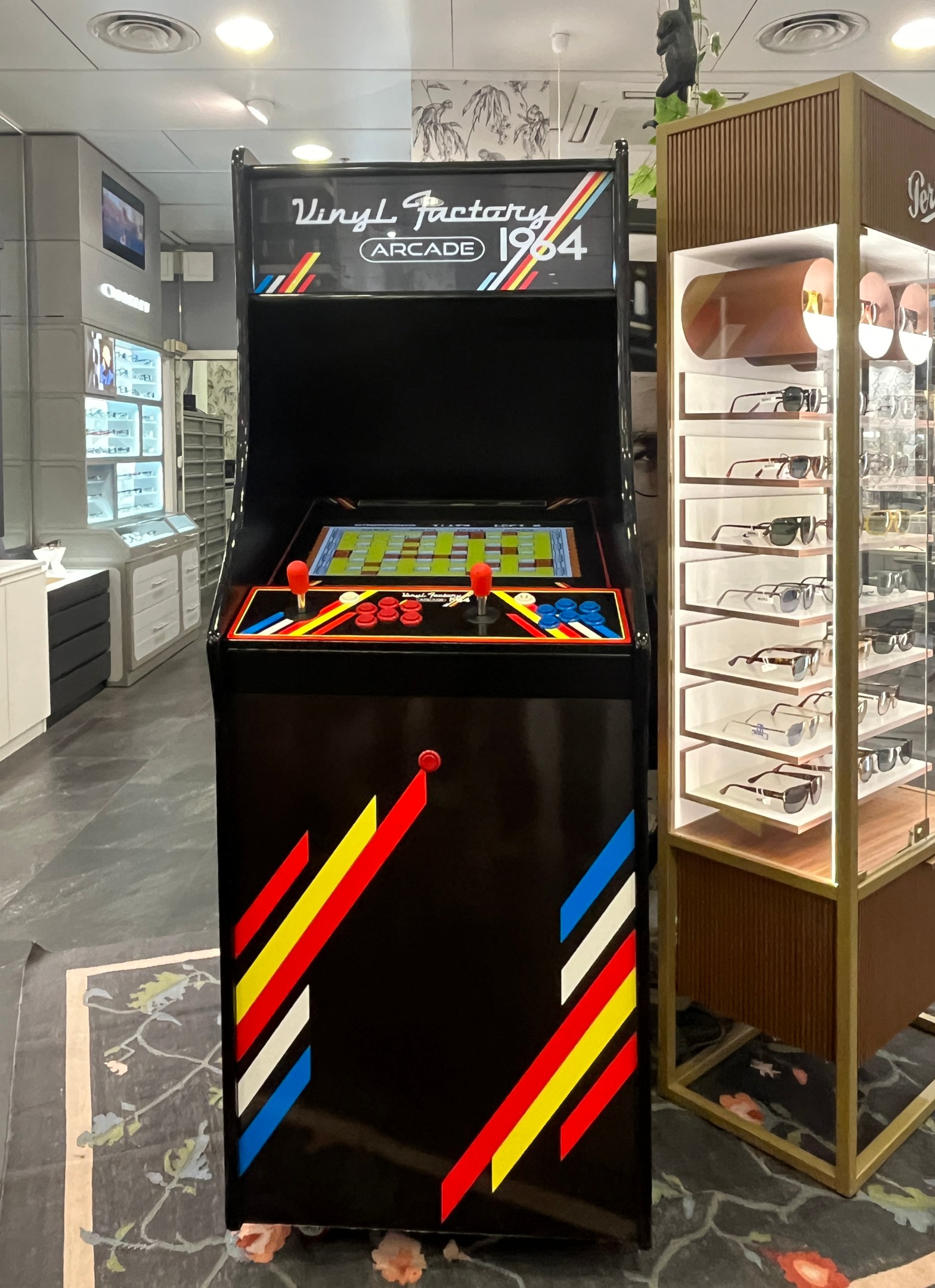 Ein Retro-Style-Laden mit einer alten Arcade-Maschine in der Mitte, einer Brillenablage rechts und Schränken, einem Fernseher und anderen Gegenständen im Hintergrund unter Deckenleuchten.