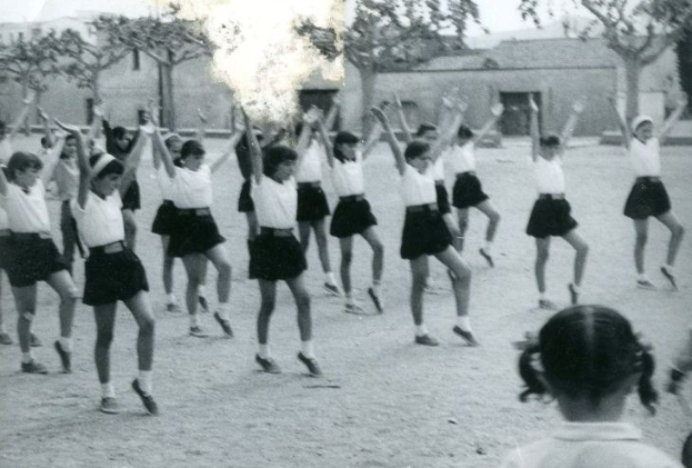 Schwarzes und weißes Bild von jungen Mädchen in Schuluniformen, die auf einer Straße tanzen, die von Gebäuden und Bäumen gesäumt ist, mit sichtbaren Füßen einer Person am Boden.