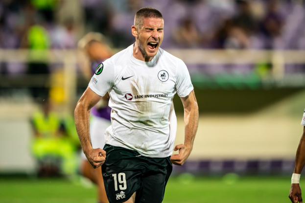 Ein Mann in weißem Hemd und schwarzen Shorts rennt auf einem Fußballfeld und feiert mit einer Gruppe im Hintergrund.