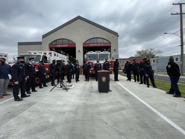 Gruppe von Menschen in Mützen neben einem Feuerwehrauto auf einem Podium während einer Feuerwache-Eröffnung, mit Gebäuden, Bäumen, Strommasten und Gras im Hintergrund unter einem bewölkten Himmel.