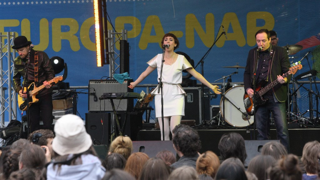 Vier Personen, die auf einer Bühne Musik machen; eine Frau in einem weißen Kleid singt in der Mitte, während zwei Männer Gitarren spielen, mit einer Menge unten und Lautsprechern gegen eine Wand im Hintergrund.