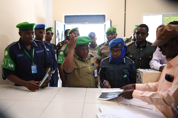 Eine Gruppe von Menschen, die um einen Tisch mit Papieren stehen, einige tragen Mützen und ID-Karten, einer hält ein Mobiltelefon, in einem Raum mit Türen und Fenstern.