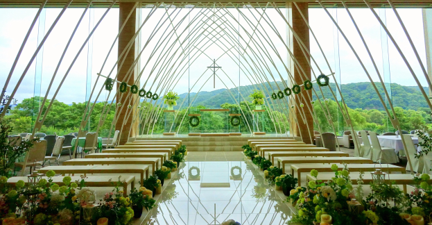 Eine Hochzeitszeremonie in einem großen, geschmückten Saal mit Tischen in Halbkreisform, umgeben von Blumenarrangements, Bäumen, Hügeln und einem klaren blauen Himmel.