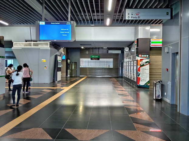 Menschen gehen durch einen Bahnhof mit einem Gepäckwagen auf der rechten Seite, Deckenleuchten, eine Uhr an der Wand und Informationsanzeigen.