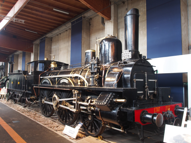 Alte Dampflokomotive in einem Museum ausgestellt, umgeben von Eisenbahnschienen, Informationsschildern und anderen Ausstellungsstücken, mit einer Wand und Deckenleuchten im Hintergrund.