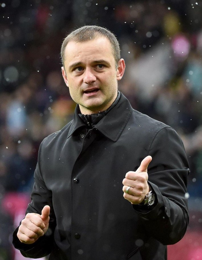 Ein Mann in einer schwarzen Jacke gibt einen Daumen hoch, trägt eine Uhr, mit einem unscharfen Hintergrund.