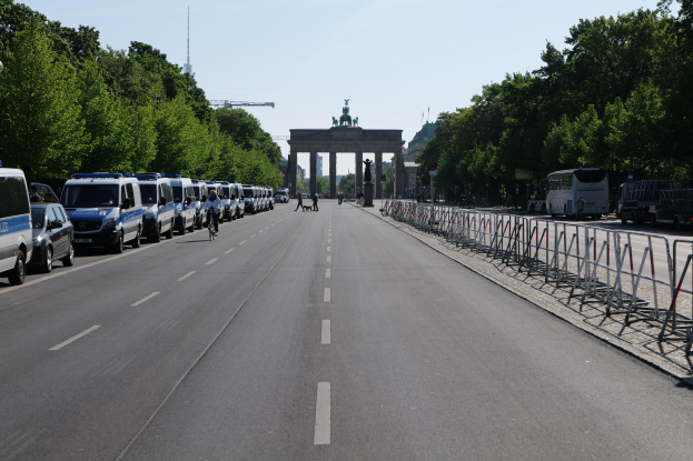 Lange Reihe von Polizeiwagen am Straßenrand vor dem Brandenburger Tor in Berlin, Deutschland, mit Menschen auf Fahrrädern und auf der Straße, Barrieren, Bäumen, einem Bogen mit Statuen im Hintergrund und dem Himmel darüber.