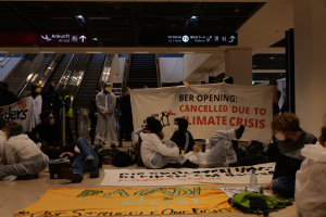 Eine Gruppe von Menschen sitzt auf dem Boden eines Flughafens und hält ein Banner in der Hand, auf dem "Berlin wegen Klimakrise gestrichen" steht. Um sie herum liegen Taschen, Flaschen und andere Gegenstände verstreut, im Hintergrund sind Rolltreppen, Tafeln mit Text und Deckenleuchten zu sehen.
