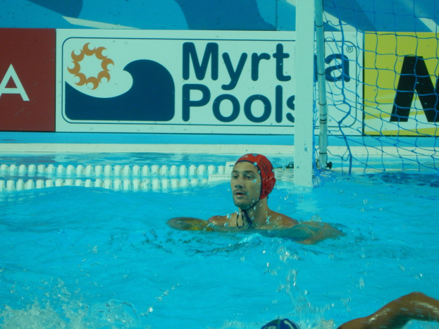 Ein Wasserballspieler mit einer Mütze, der in einem Pool schwimmt, mit einem Netz auf der rechten Seite und einem Werbeplakat im Hintergrund.