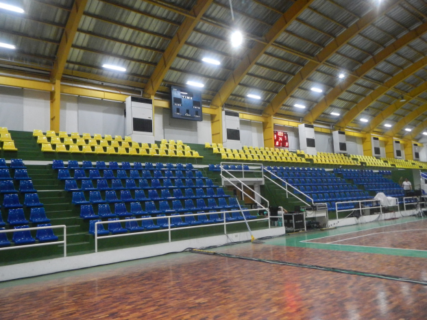 Großer Hallensportplatz mit Holzboden, blauen und gelben Sitzplätzen, Geländern, Treppen zum Sitzbereich, wandmontierten Bildschirmen, Deckenleuchten und einigen Personen.