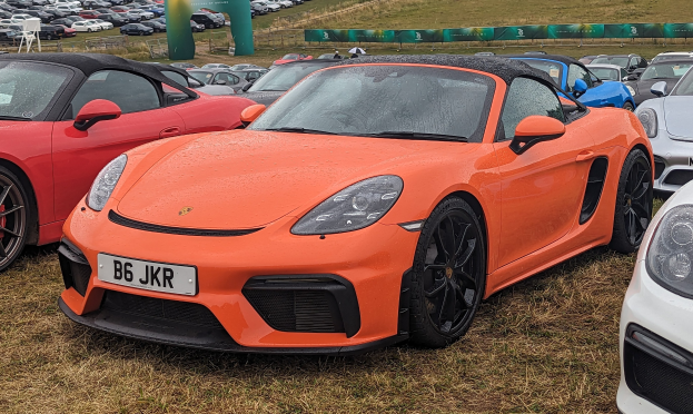 Porsche 718 Boxster GTS auf dem Goodwood Festival of Speed, umgeben von anderen Autos auf Gras, mit einem Banner, Bäumen und einem klaren blauen Himmel im Hintergrund.
