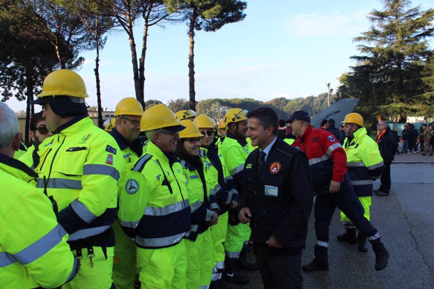 Gruppe von Einsatzkräften in gelben Westen und Helmen auf einer Straße mit Bäumen und einem klaren blauen Himmel.