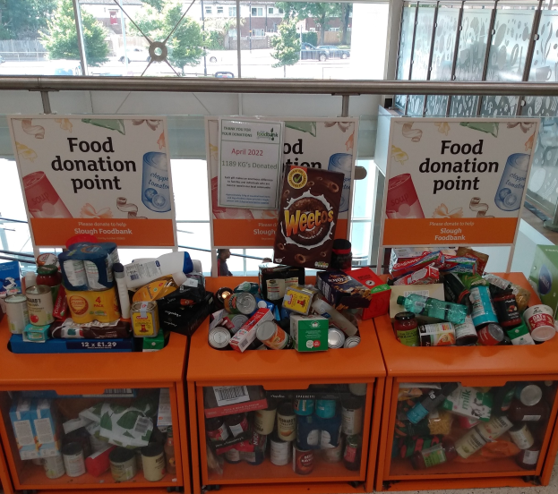 Orangene Schränke mit Lebensmitteln wie Flaschen, Dosen und Kartons an einer Spendenstelle, mit Informationsschildern und einem Blick durch Glasfenster auf Fahrzeuge, Bäume, Gebäude und Himmel.