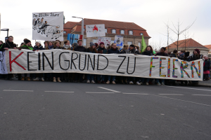 Demonstranten mit einem Transparent mit der Aufschrift 'Kein Grund zur Feier' gegen deutsche Sparmaßnahmen, mit Straßeninfrastruktur und Gebäuden im Hintergrund bei klarem Himmel.