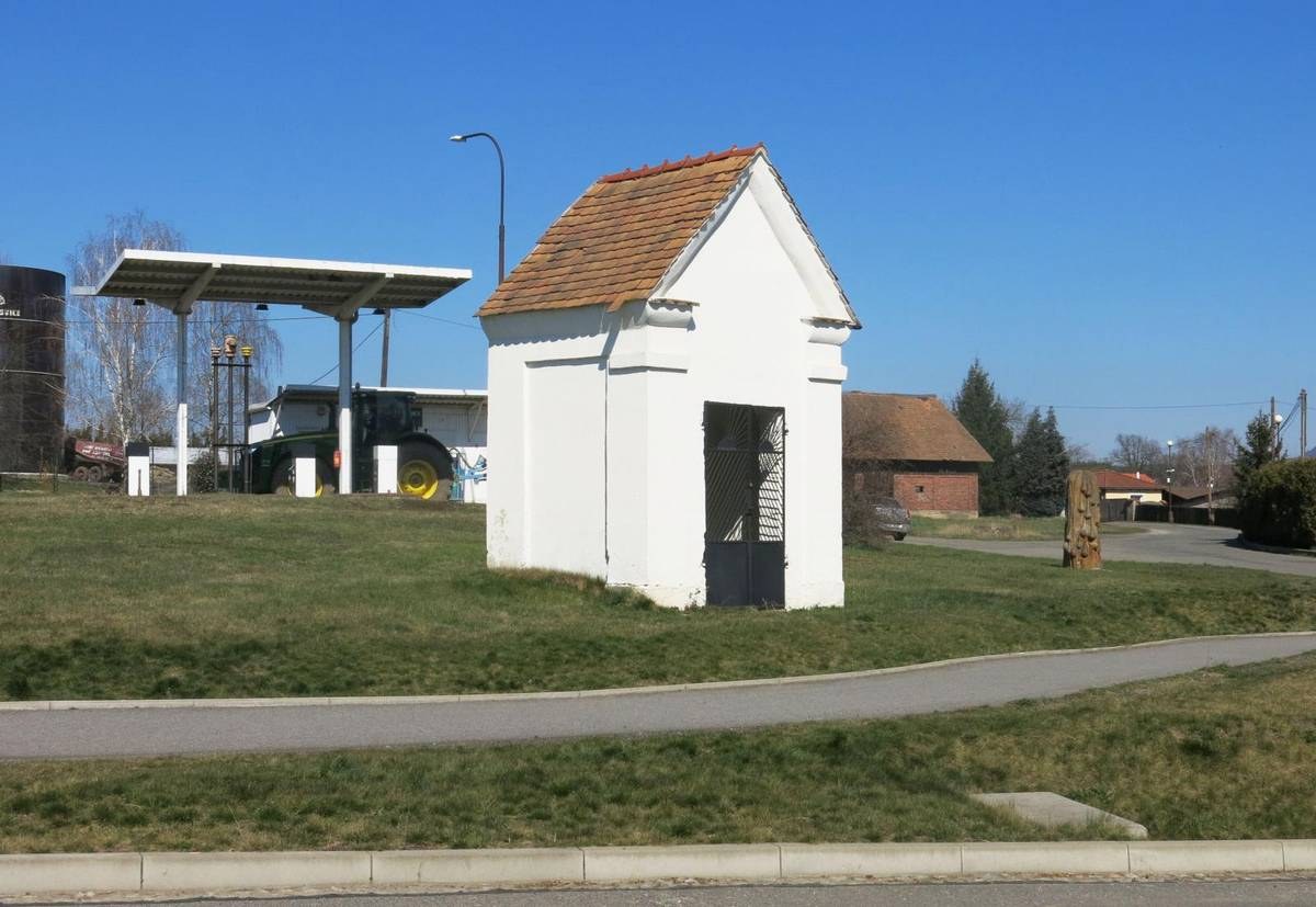 Kleines weißes Gebäude mit einem roten Dach, die erste Tankstelle in Deutschland, neben einer Straße mit Gras, Bäumen, Strommasten und Häusern im Hintergrund unter einem klaren blauen Himmel.