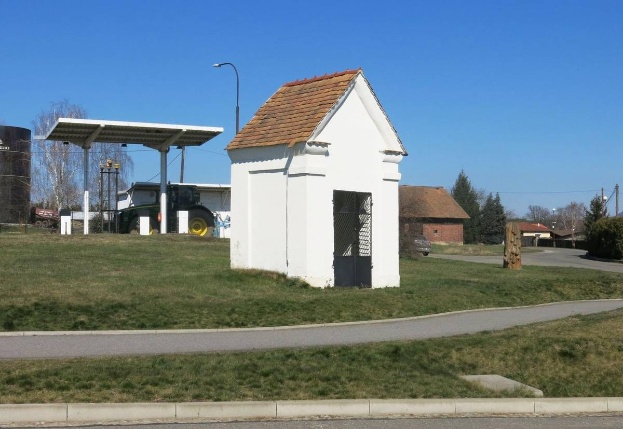 Kleines weißes Gebäude mit einem roten Dach, die erste Tankstelle in Deutschland, neben einer Straße mit Gras, Bäumen, Strommasten und Häusern im Hintergrund unter einem klaren blauen Himmel.