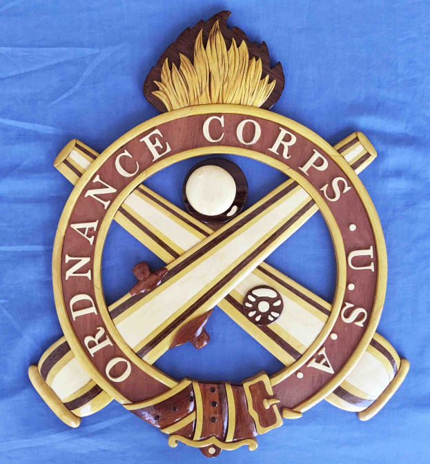 Ein hölzernes Schild mit "Ordnance Corps" in schwarzer Schrift, das ein geschnitztes Design mit einem weißen Stern auf einem navyblauen Hintergrund und einem goldenen Rand zeigt, auf blauem Stoff montiert.